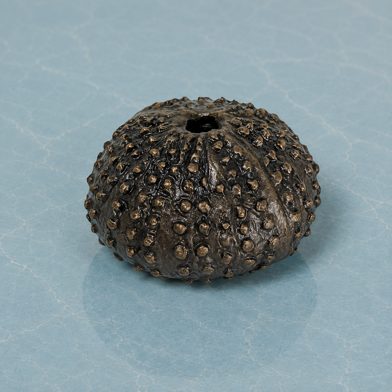 A small sea urchin bronze sculpture with a textured surface sat on a light blue marble background.
