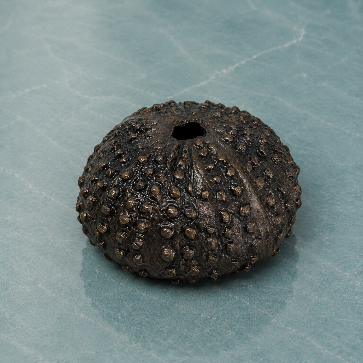 Textured solid bronze sea urchin miniature sculpture sat on a light blue marble surface.