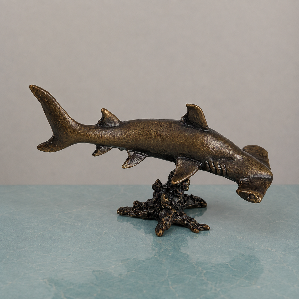 
                  
                    Bronze hammerhead shark figurine mounted on a textured coral-like base, displayed against a neutral gray background and light blue surface.
                  
                